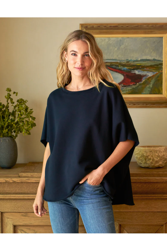 Woman wearing a black oversized shirt and blue jeans standing in a room with a painting and plant in the background.
