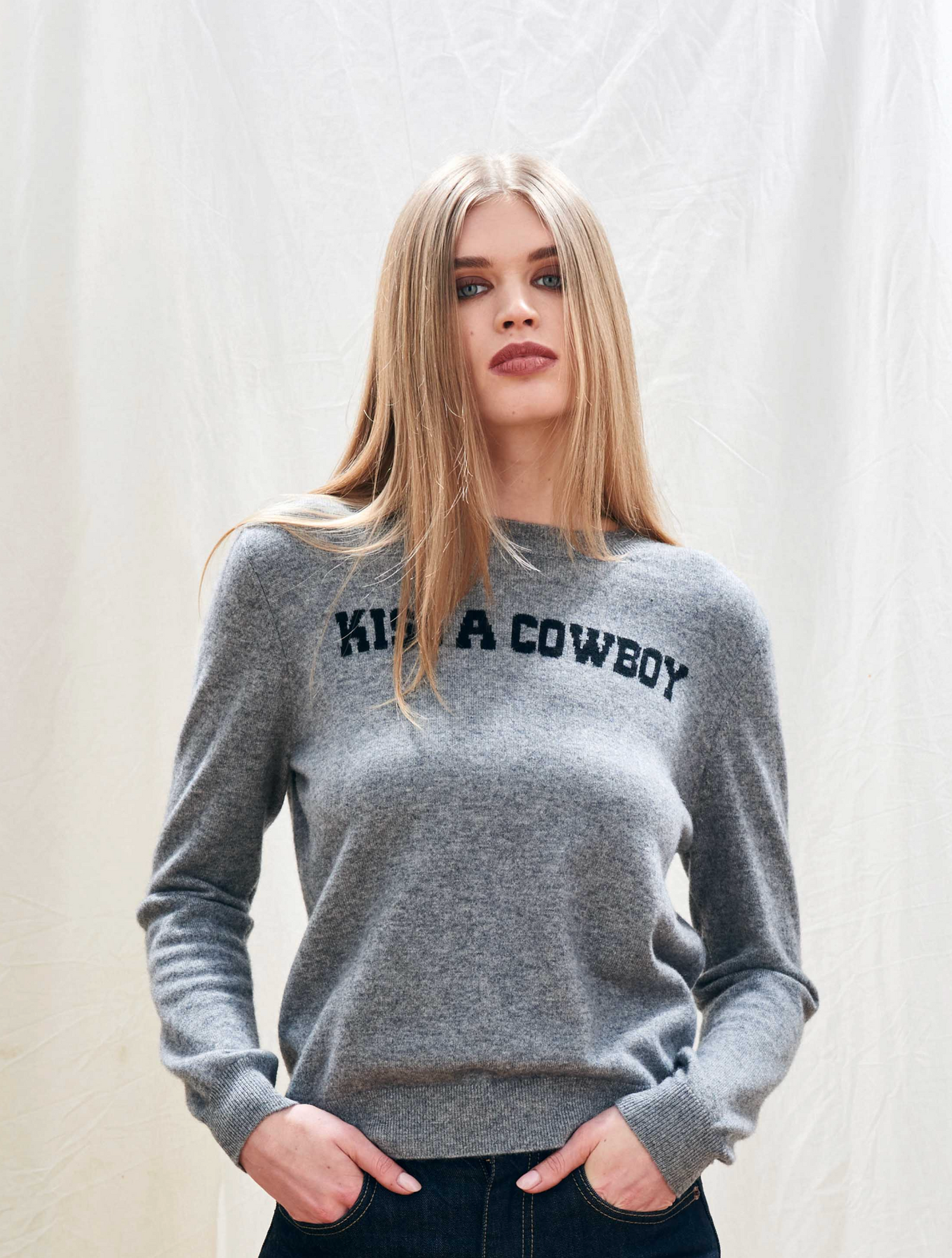 Woman wearing a gray sweater with 'Kiss a Cowboy' text on a plain background