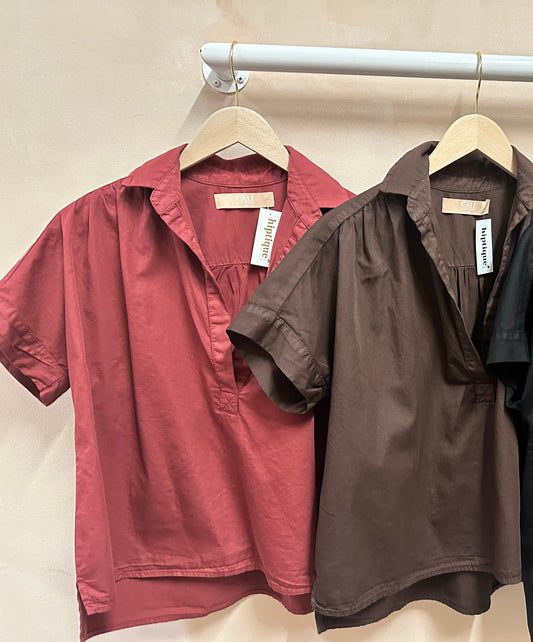 Three short-sleeve shirts in red, brown, and black hanging on a rack against a beige wall.