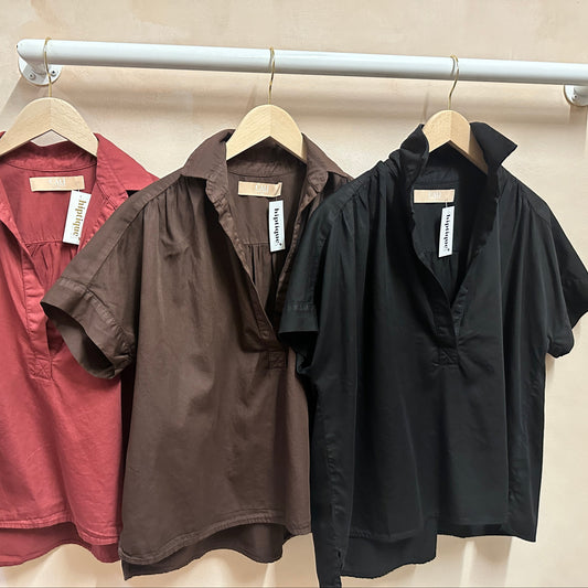 Three short-sleeve shirts in red, brown, and black hanging on a rack against a beige wall.