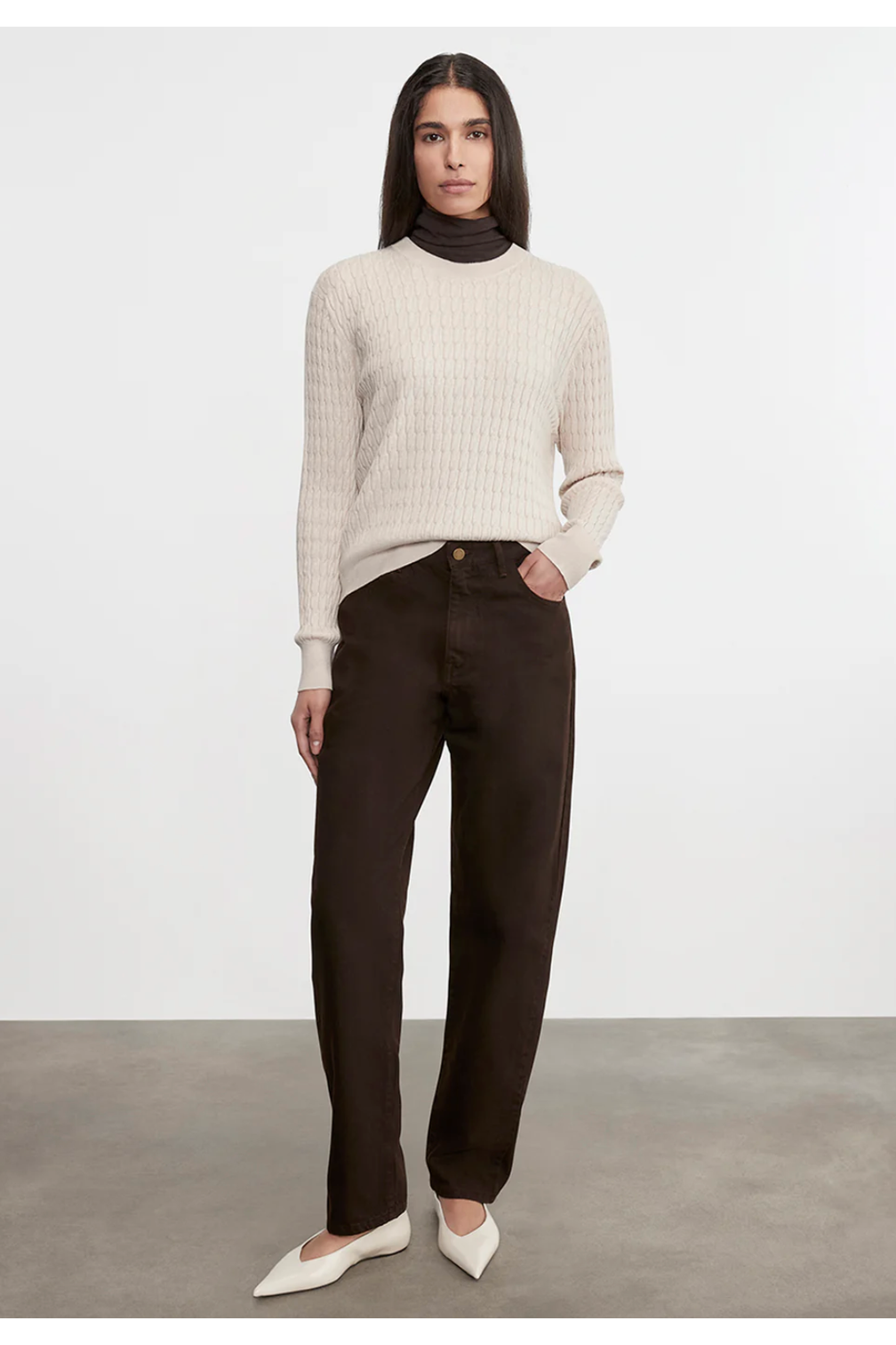 Woman wearing a beige sweater and dark pants against a white background