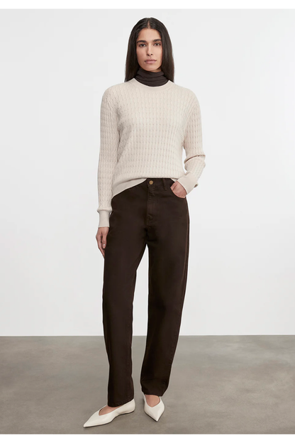 Woman wearing a beige sweater and dark pants against a white background
