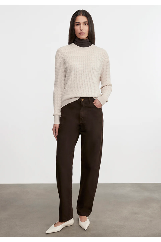 Woman wearing a beige sweater and dark pants against a white background