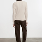 Person wearing a white ribbed sweater and dark pants against a plain background