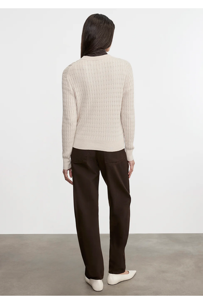 Person wearing a white ribbed sweater and dark pants against a plain background