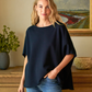 Woman wearing a black oversized shirt and blue jeans standing in a room with a painting and plant in the background.