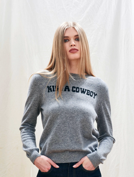 Woman wearing a gray sweater with 'Kiss a Cowboy' text on a plain background