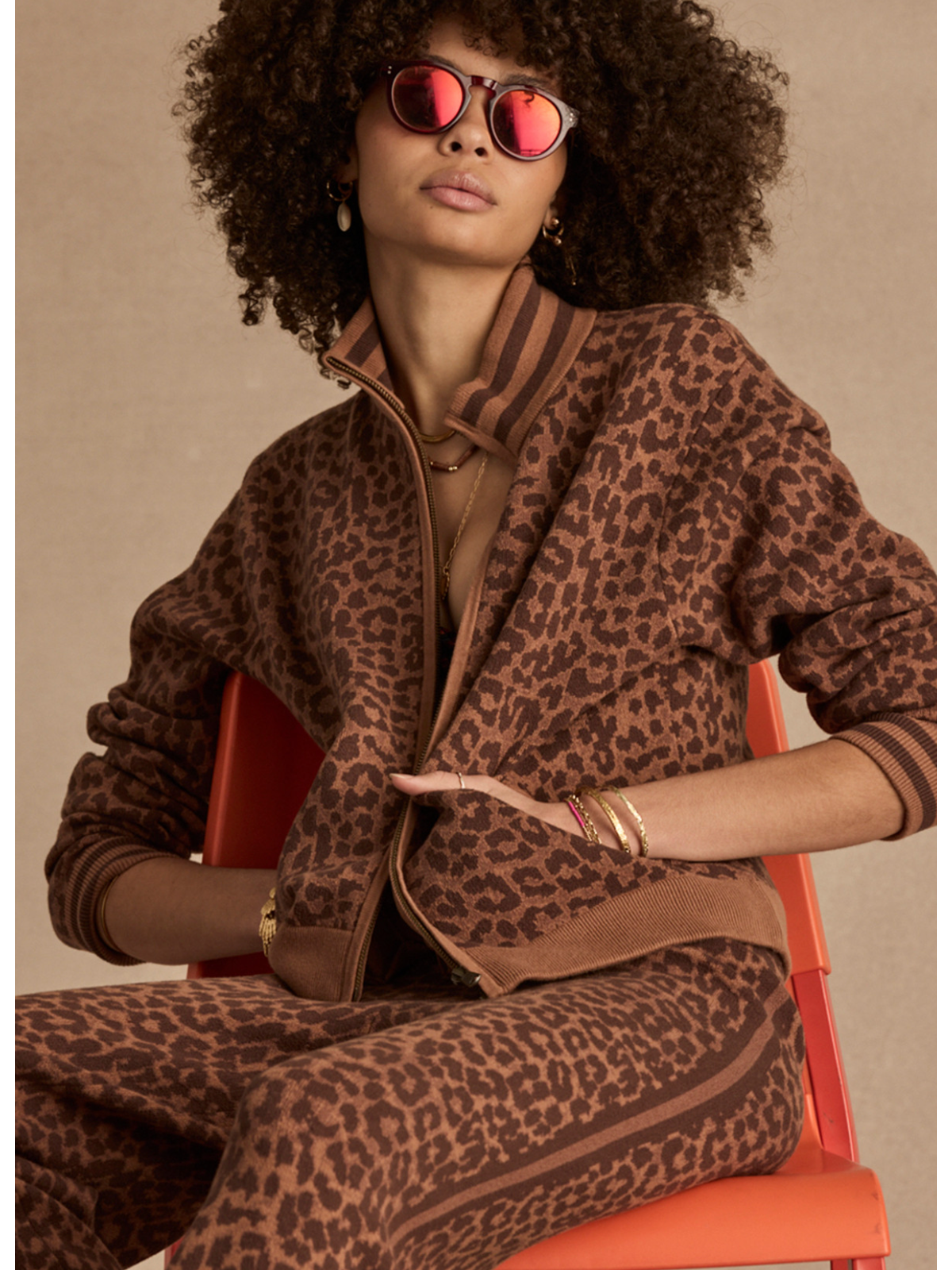 Woman wearing a leopard print outfit with sunglasses on an orange chair against a beige background